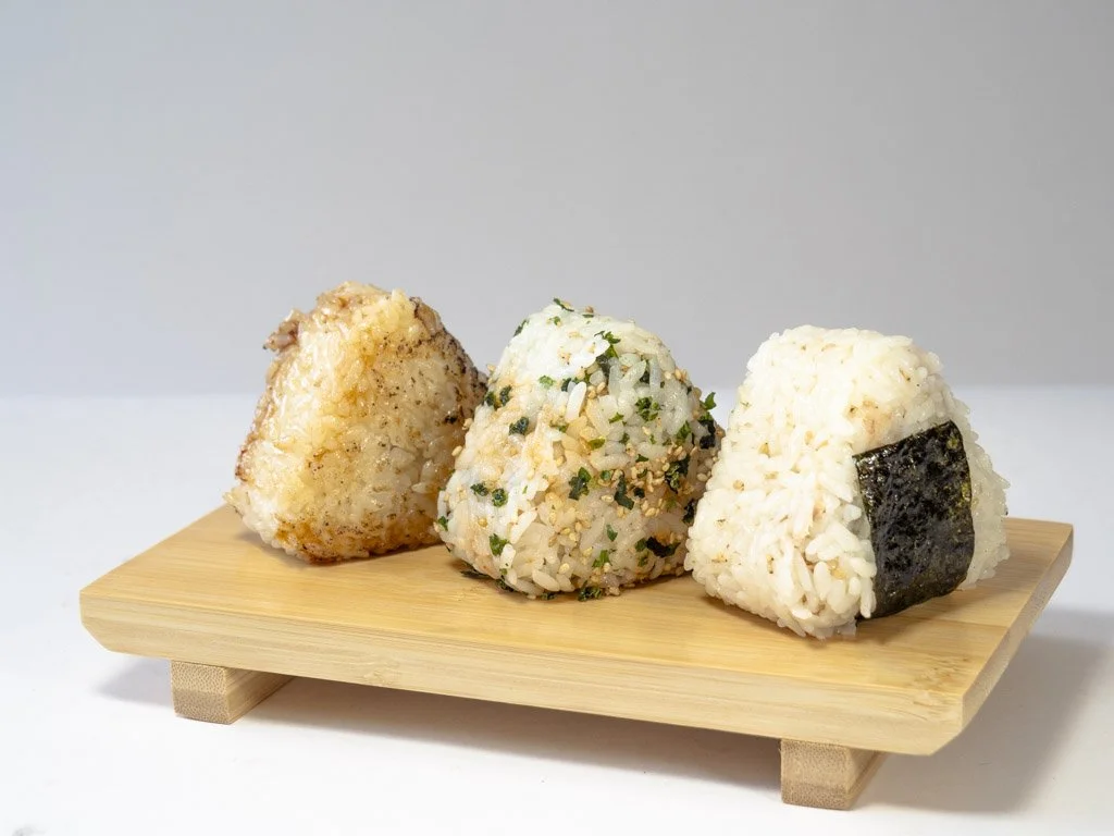 Three rice balls on a small wooden tray, each with different toppings or fillings.