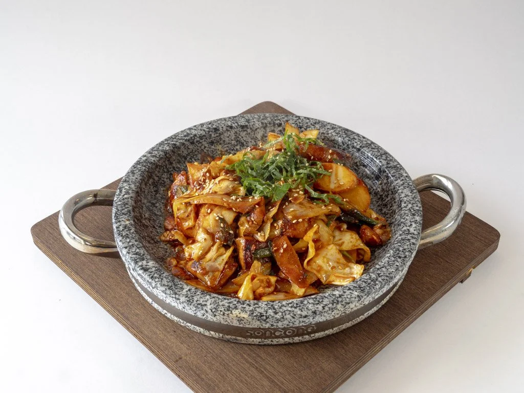 A bowl of spicy Korean-style stir-fried vegetables with cabbage, carrots, and green garnish, served in a gray stone bowl on a wooden tray.