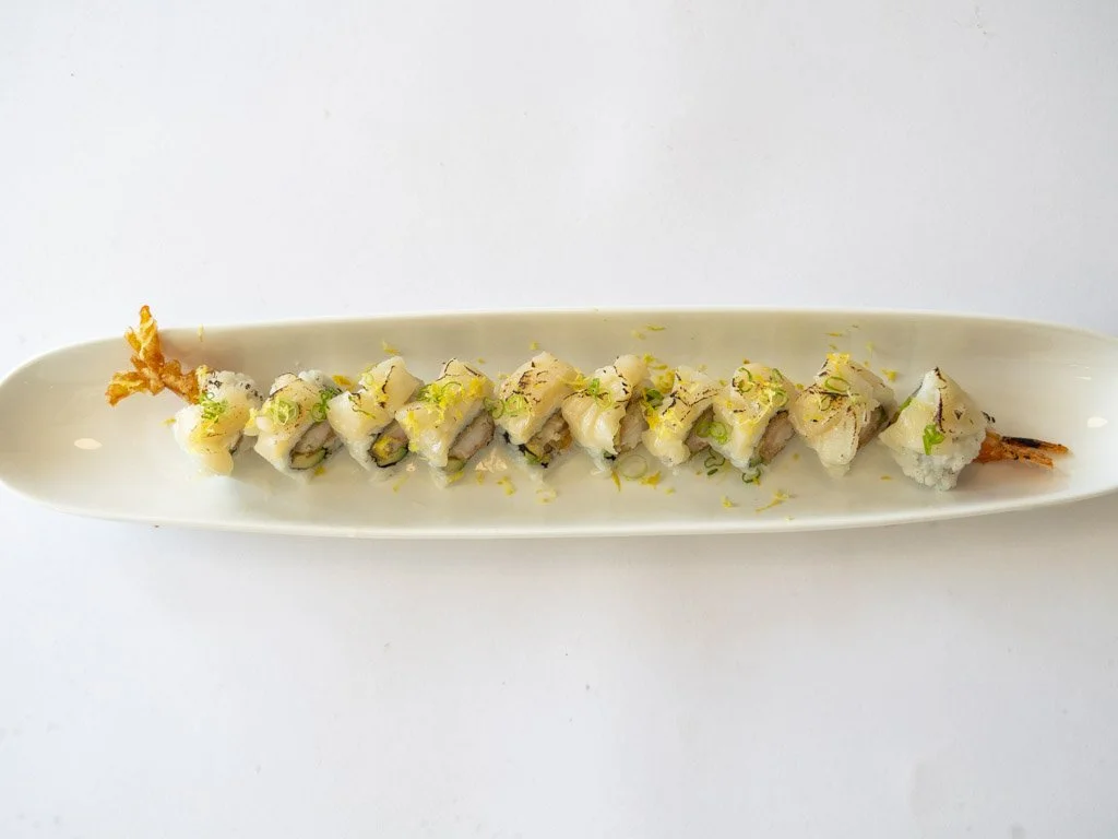 A long white plate with pieces of sushi roll topped with chopped green onions and lemon zest, garnished with small orange fish roe, on a white background.