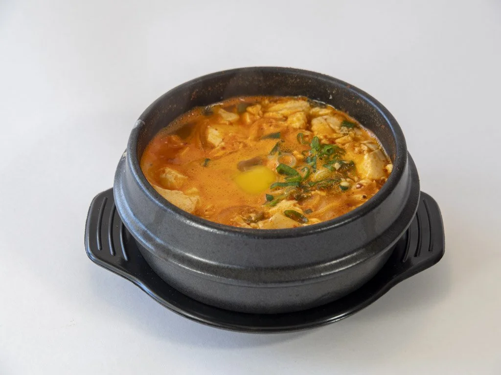 A black ceramic bowl filled with spicy Korean stew, showing a runny yellow egg yolk, vegetables, and green onions, placed on a black saucer against a white background.