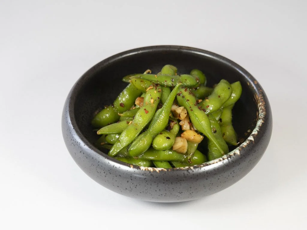 A black ceramic bowl filled with sautéed green edamame beans topped with chopped garlic and red pepper flakes.