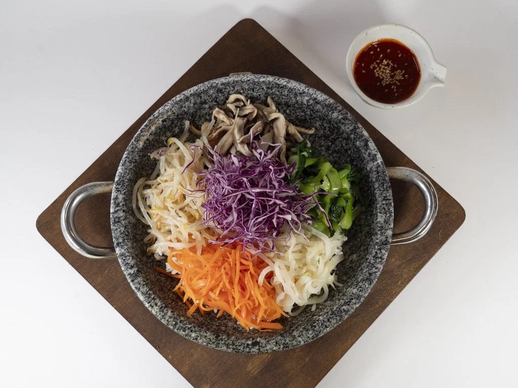 A bowl of various colorful sliced vegetables including purple cabbage, green onions, shredded carrots, shredded white radish, and mushrooms, with a small cup of dipping sauce on the side.