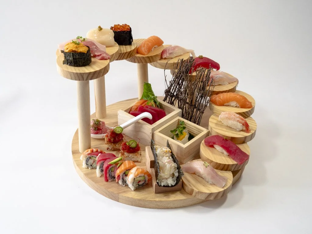 A wooden display stand with various types of sushi including nigiri, sashimi, and rolls, presented on small round platforms, with some garnishes and decorative elements.