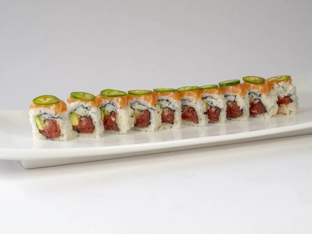 Sushi roll with salmon, avocado, and raw fish, topped with sliced jalapeño peppers, arranged on a white rectangular plate.