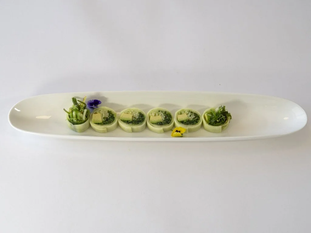 A long white plate holds six finely sliced cucumber rolls filled with a green mixture and garnished with edible flowers and small greens.