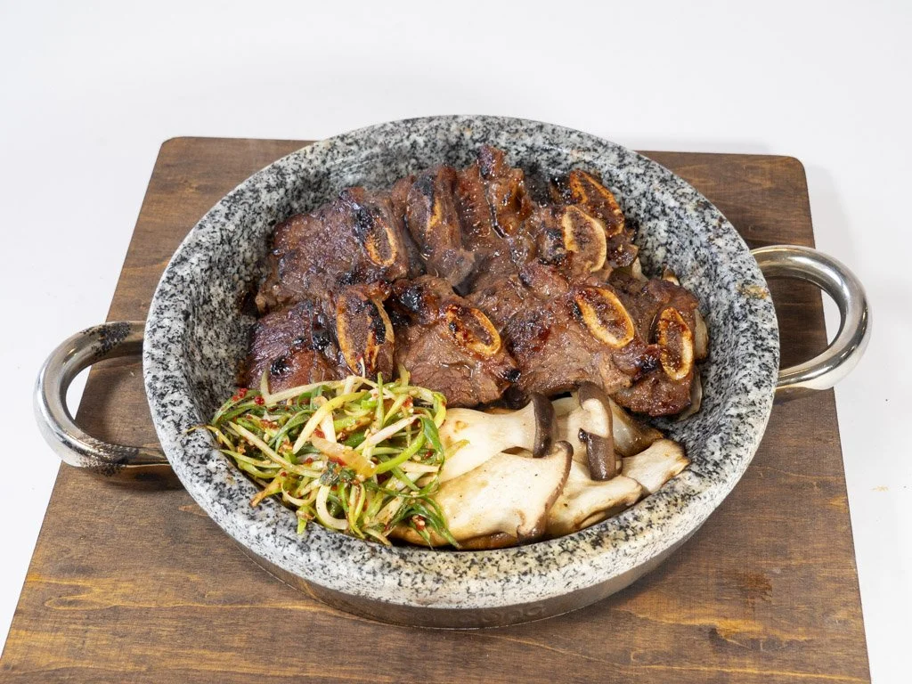 A stone bowl with meat, sautéed vegetables, and mushrooms on a wooden serving board.