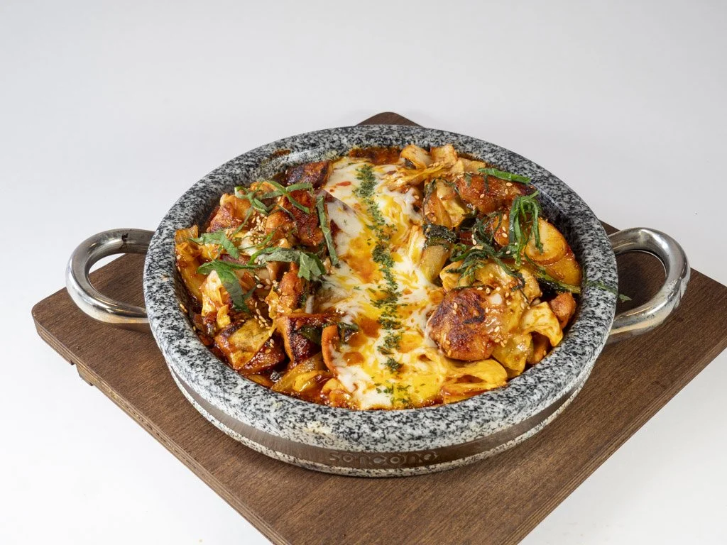 A skillet of baked pasta with melted cheese, vegetables, and herbs, served on a wooden board.