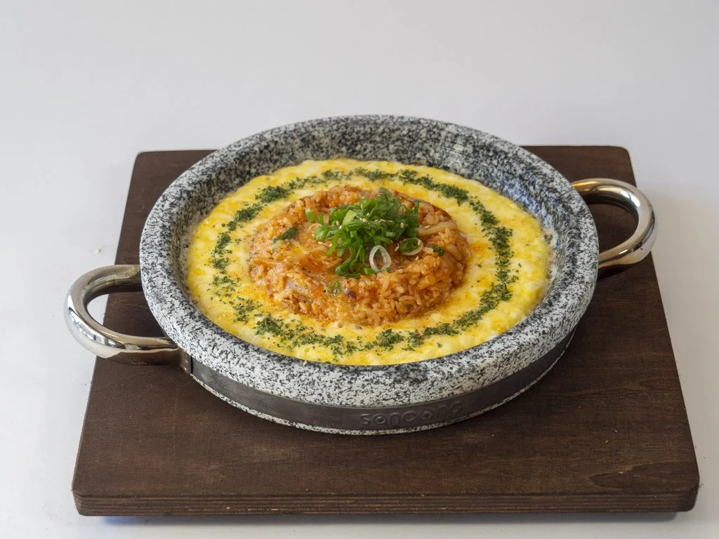 A stone bowl of spicy fried rice topped with melted cheese, garnished with chopped green onions, served on a wooden board.