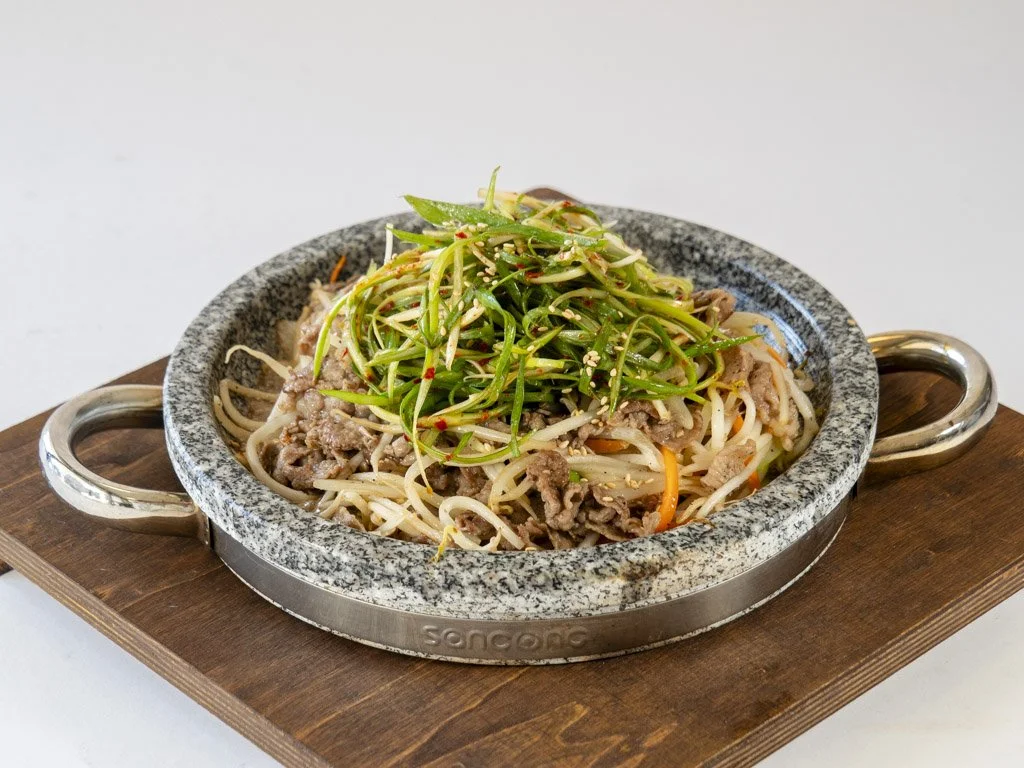 A bowl of Asian stir-fried noodles with vegetables and beef, topped with shredded green vegetables and sesame seeds, served on a stone hot plate on a wooden tray.