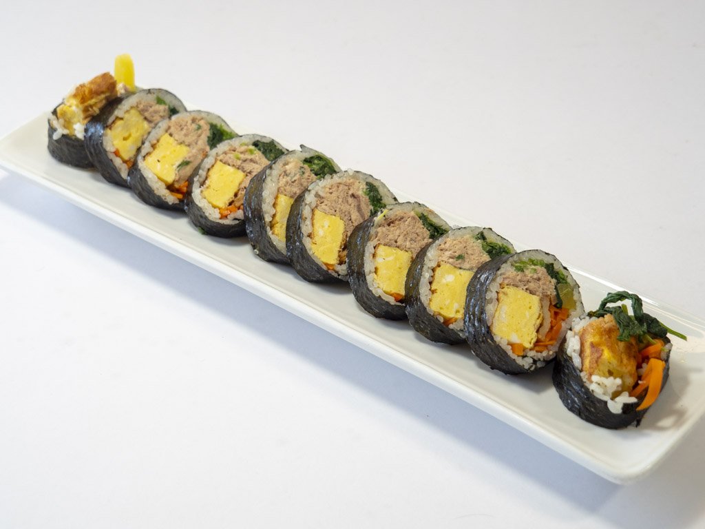 Japanese sushi rolls on a white rectangular plate, filled with rice, seaweed, egg, tuna, and vegetables.