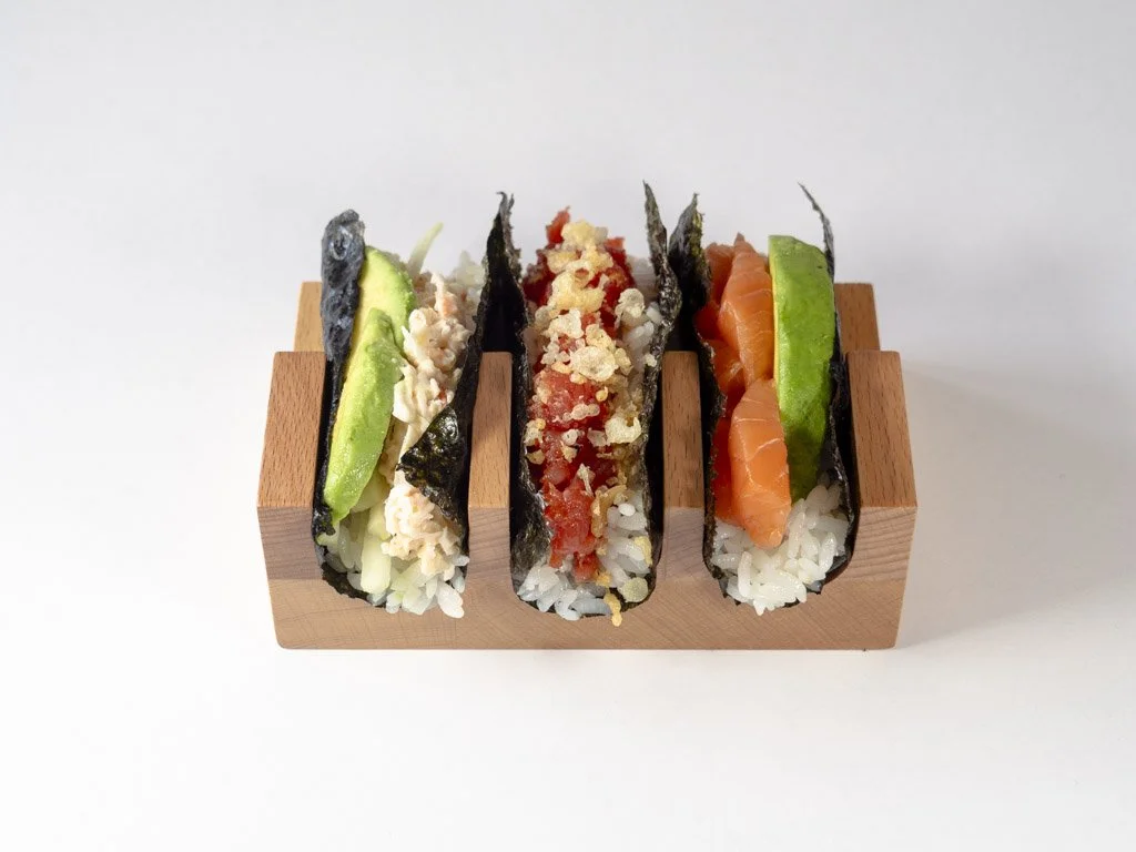 Three sushi tacos with rice, seaweed, avocado, crab meat, fish roe, and salmon, served on a wooden block.