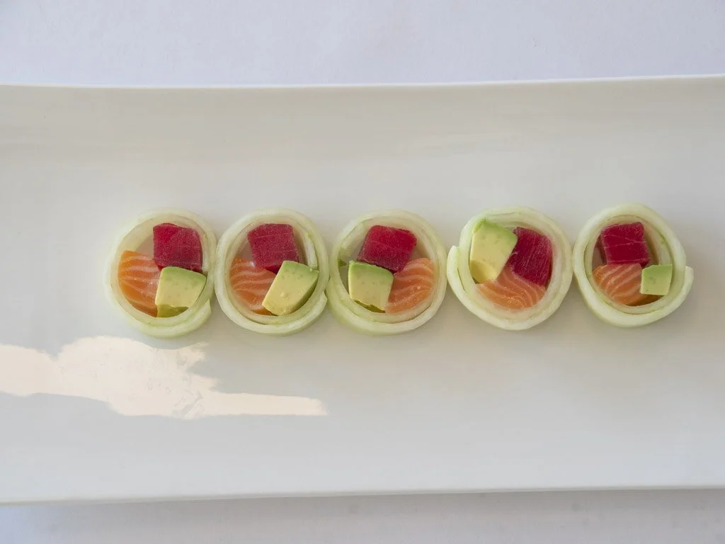 Five pieces of sushi with salmon, avocado, tuna, and cucumber, arranged on a white rectangular plate.