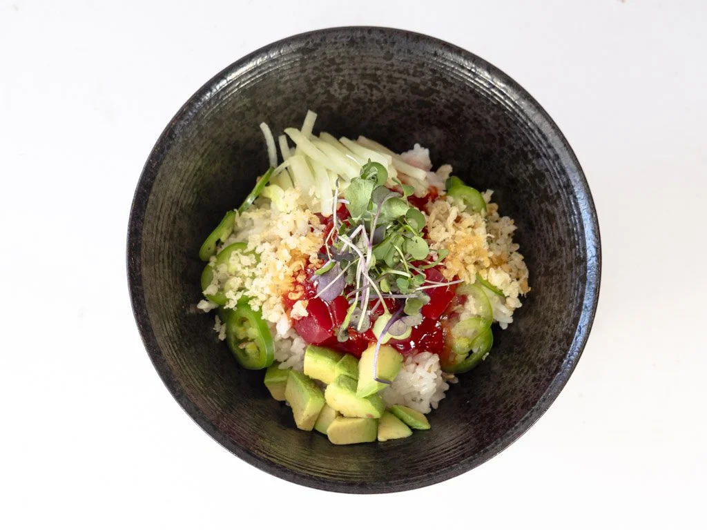 A bowl of sushi rice topped with avocado, chopped green peppers, shredded cheese, microgreens, and drizzled with red sauce.