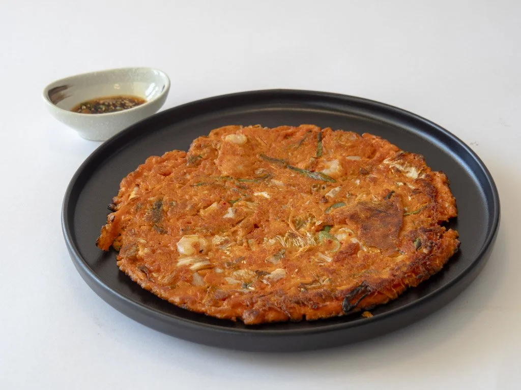 A Korean kimchi pancake on a black plate with a small side dish of dipping sauce.