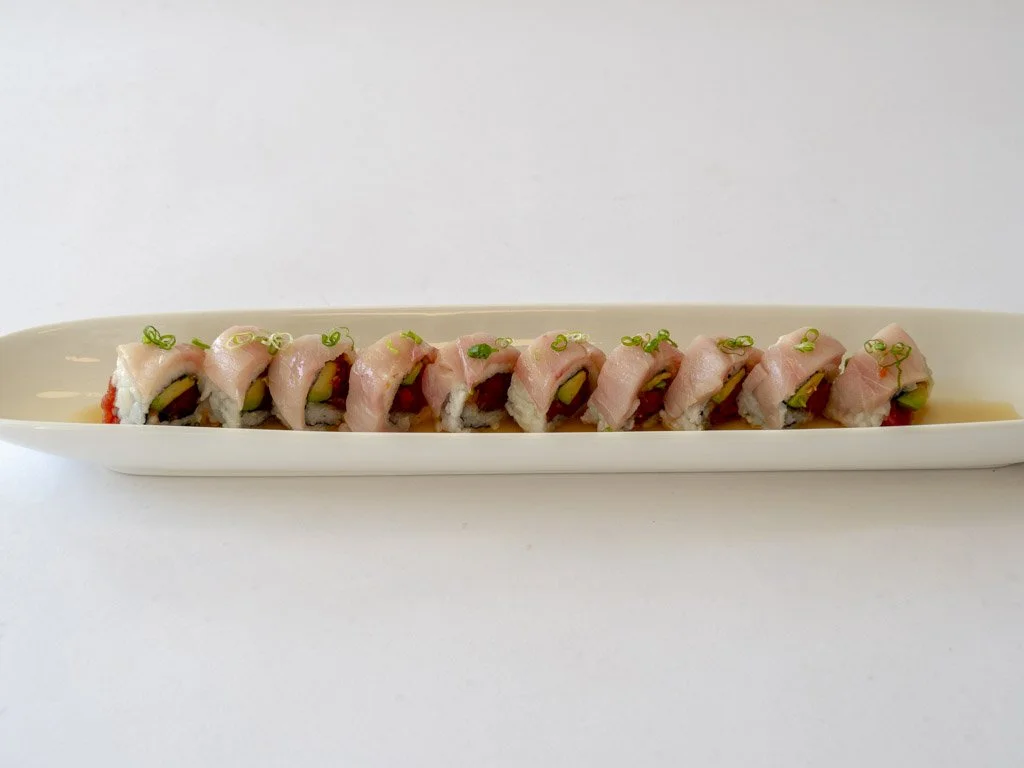A long white platter with a row of sushi rolls topped with slices of fish and garnished with green onions, placed against a plain white background.