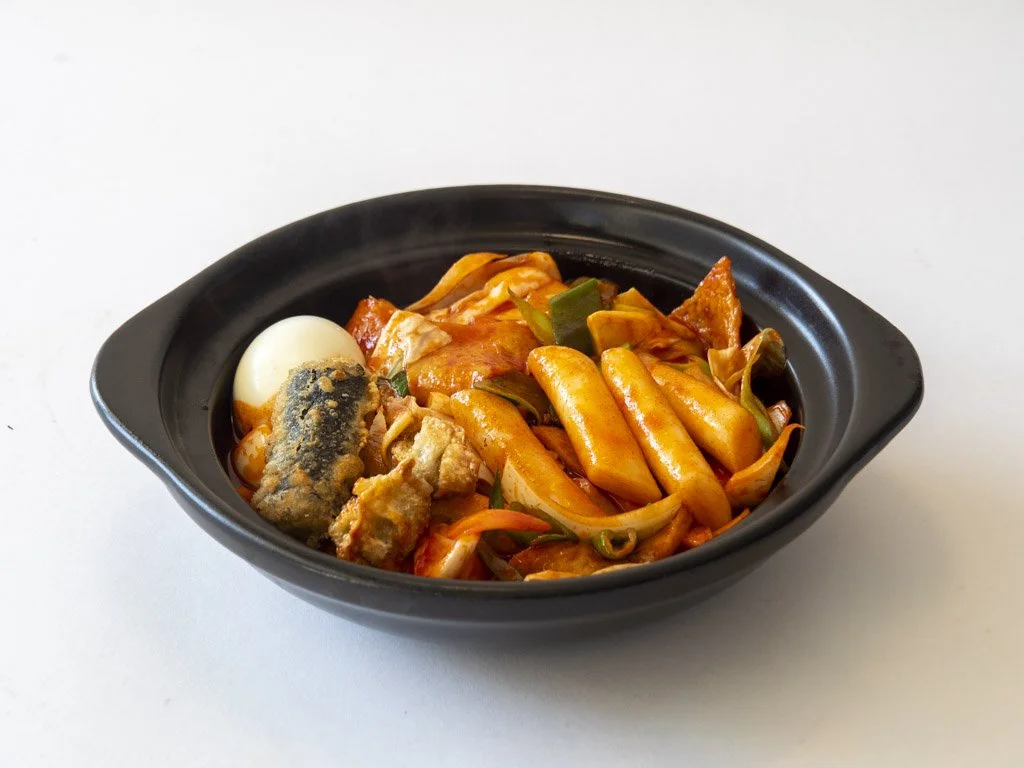 Black bowl containing Korean tteokbokki with rice cakes, fish cake, boiled egg, and vegetables in spicy sauce.