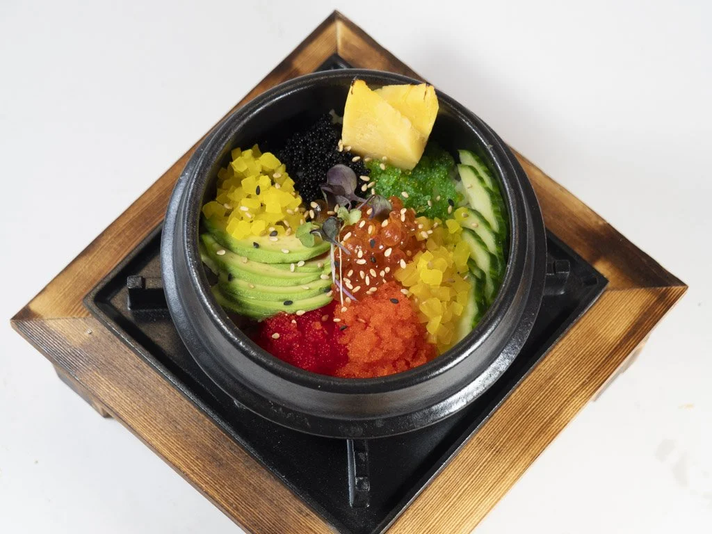 A bowl of poke with sliced avocado, pineapple, cucumber, fish roe, and other colorful ingredients on a wooden tray.