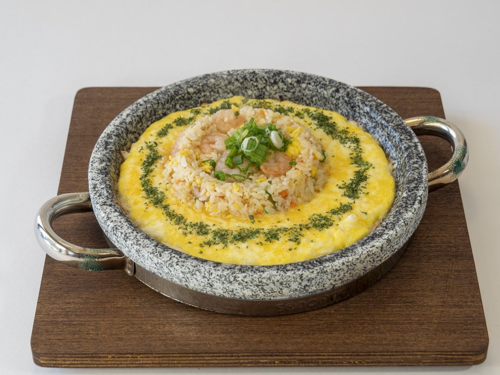 Shrimp fried rice served in a sizzling stone bowl with a fried egg and green onions on top, garnished with herbs.