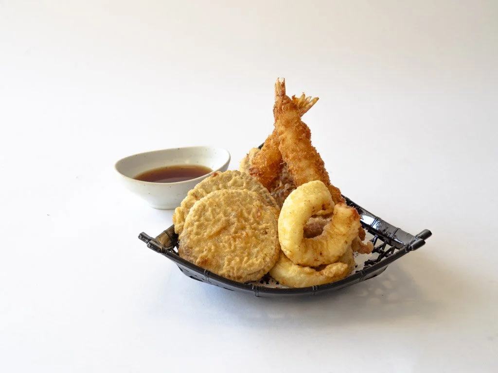 A serving basket of fried seafood, including shrimp, onion rings, and fish fillets, with a small bowl of dipping sauce on a white background.