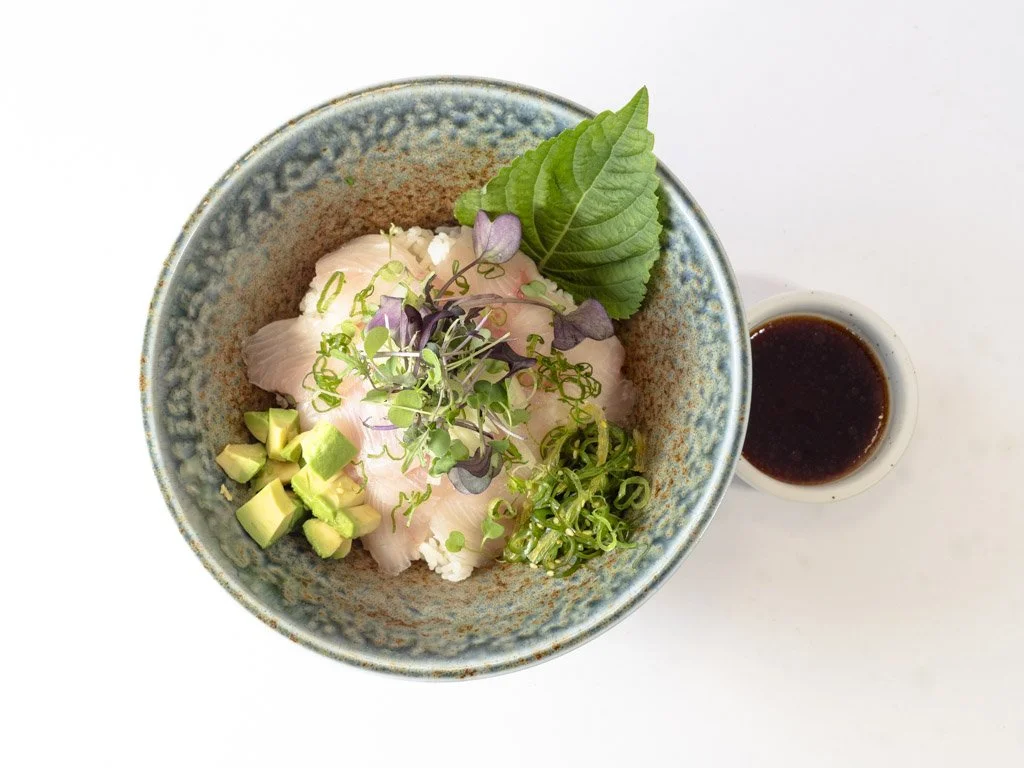 Bowl of sushi with raw fish, chopped avocado, green onions, microgreens, and leaf garnish, accompanied by a small cup of soy sauce.