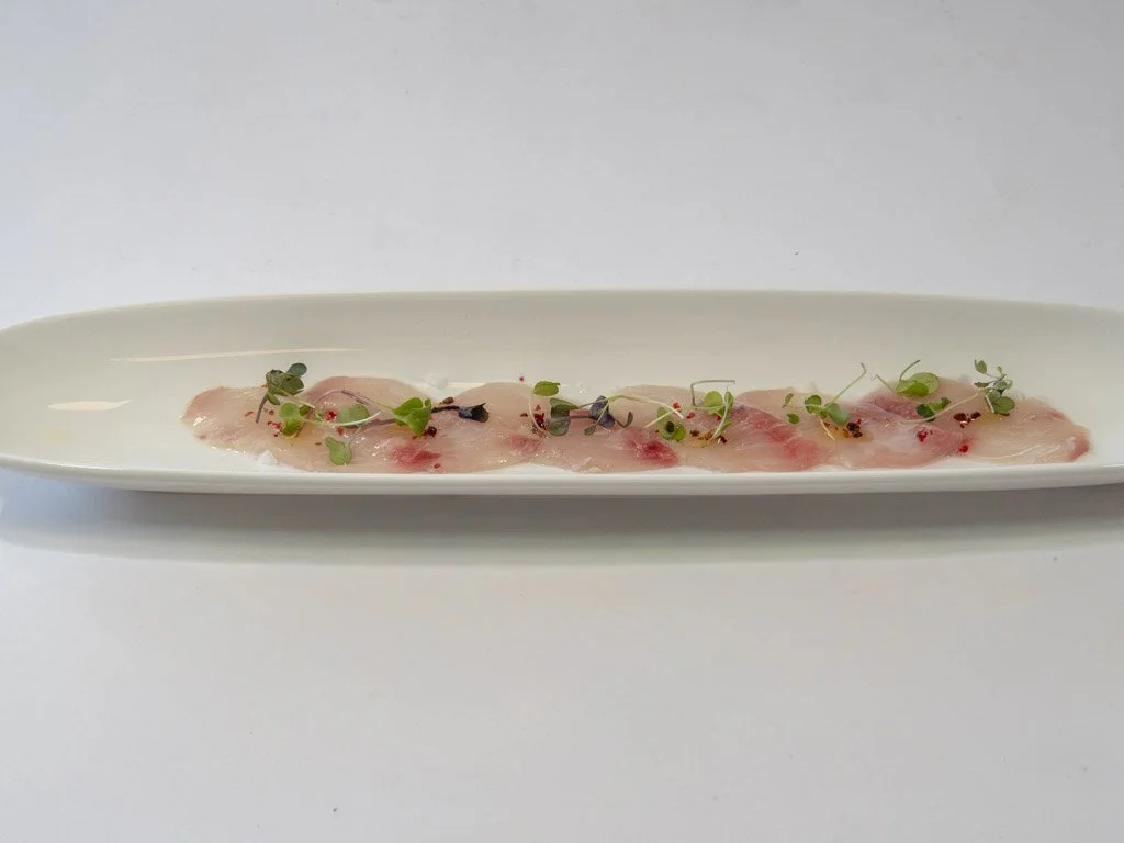 Thinly sliced raw fish arranged on a long white plate, garnished with microgreens and small red and black seasoning particles.