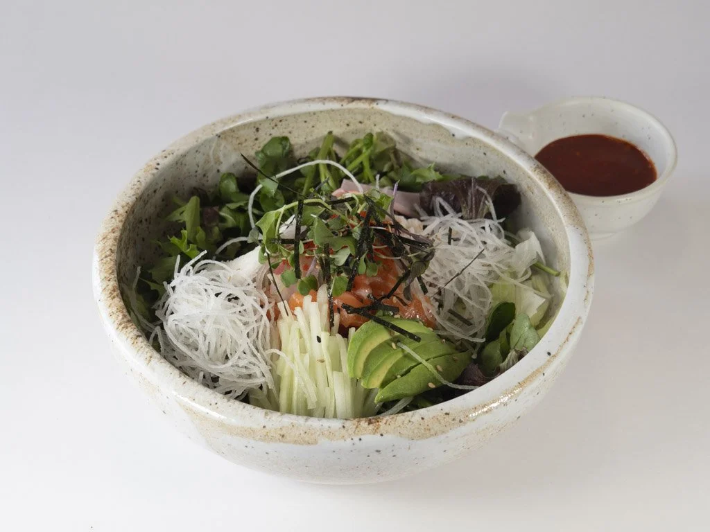 Bowl of Asian salad with mixed greens, rice noodles, sliced avocado, and shredded vegetables, served with a side of red dipping sauce.