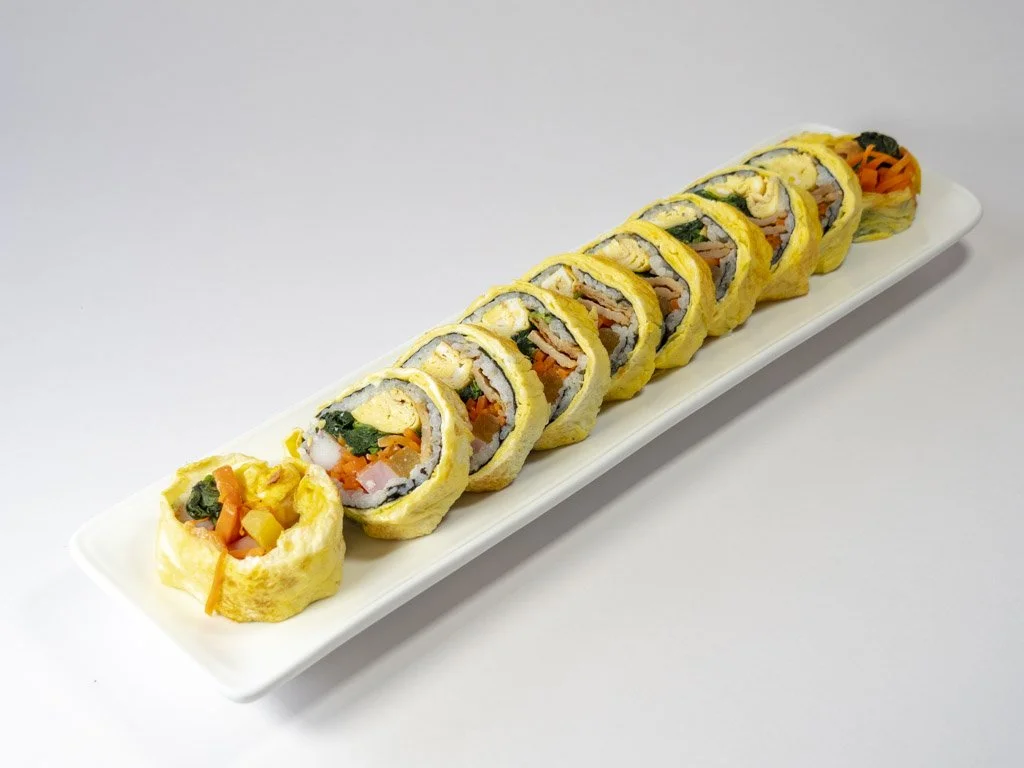 A long white plate with eight sushi rolls and one sushi roll topped with vegetables, all arranged in a row on a plain white background.