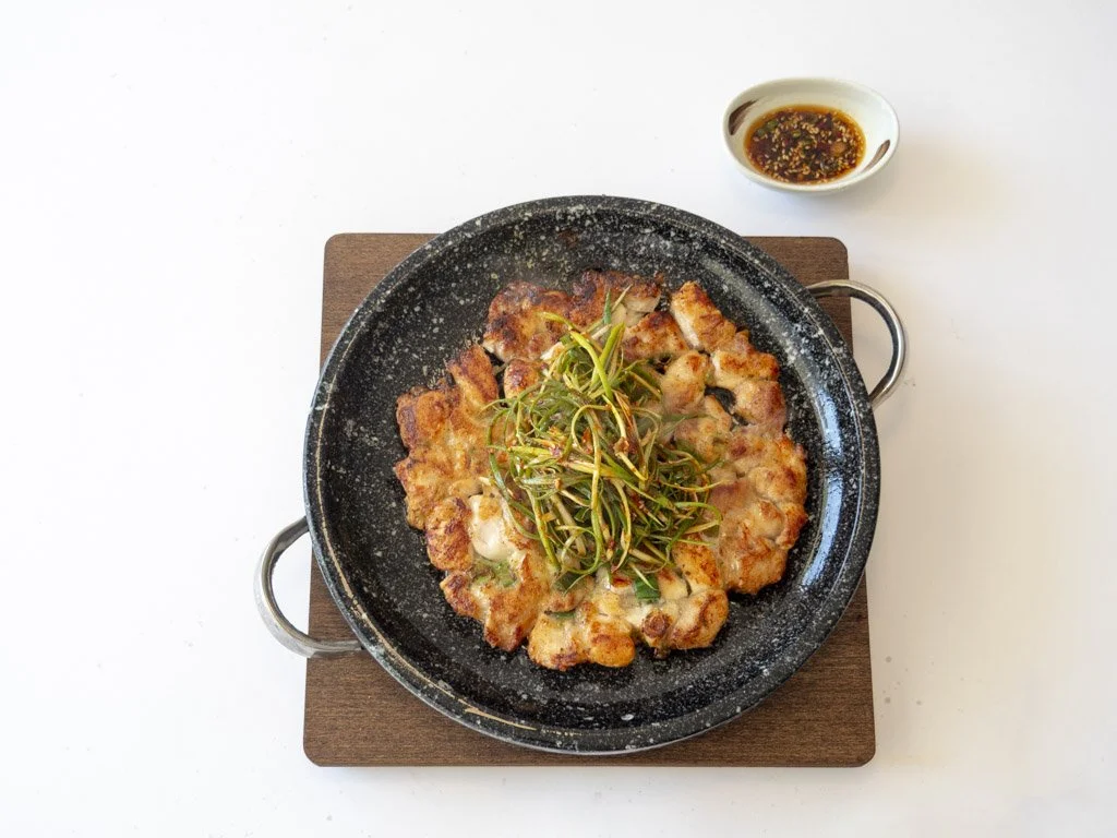 A sizzling plate of grilled chicken with green herbs on top, served with a small bowl of soy sauce-based dipping sauce placed on a wooden board.