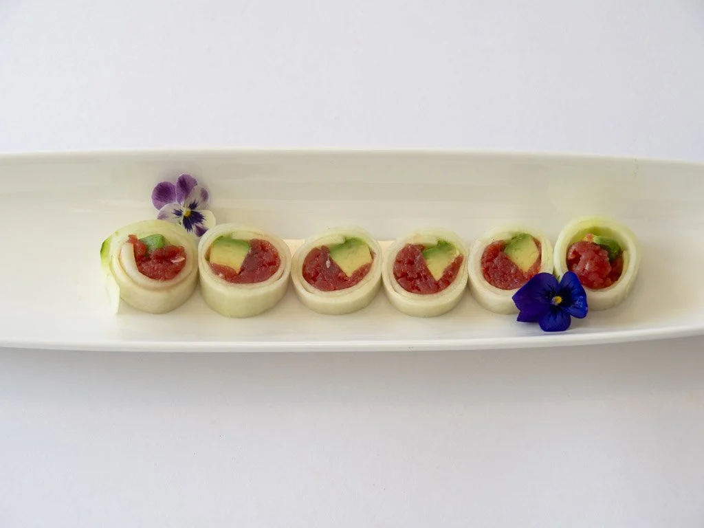Six cucumber rolls with red filling, garnished with purple flowers on a long white rectangular plate.