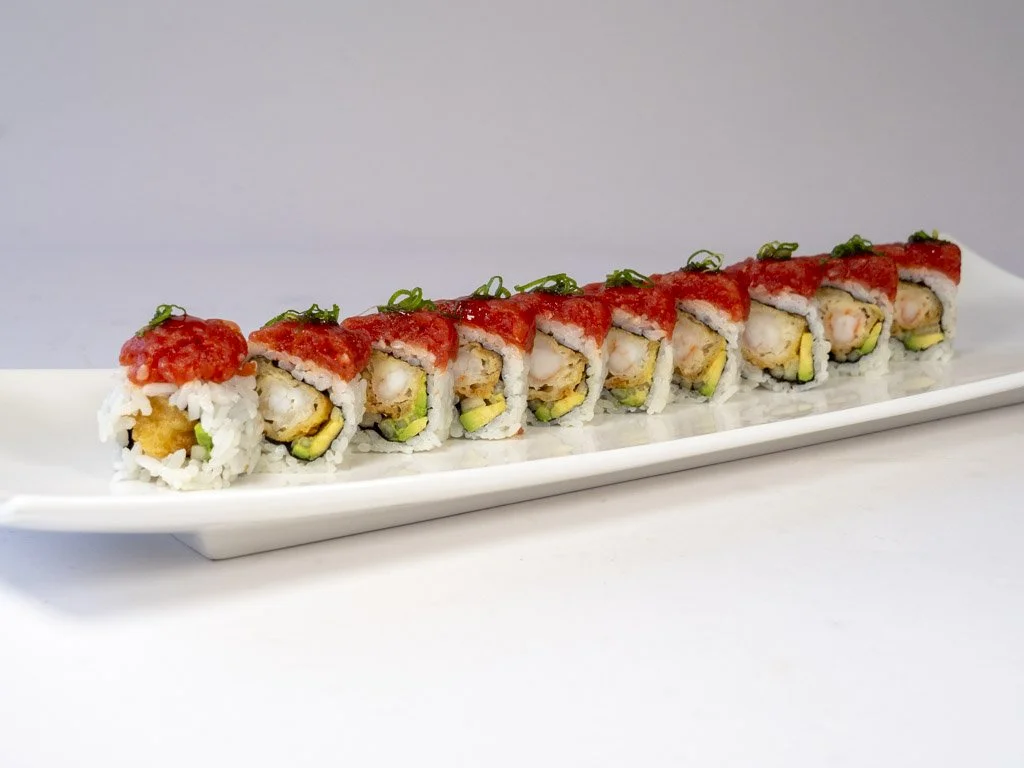 A long white rectangular plate holding a row of sushi rolls topped with red sauce and green garnishes.