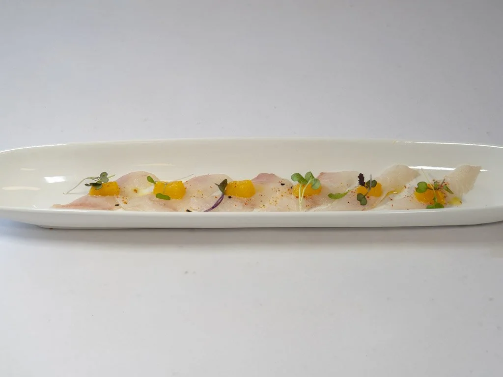 Thinly sliced fish garnished with small edible greens, microgreens, and tiny yellow fruit or vegetable pieces, served on a long, white, narrow plate.