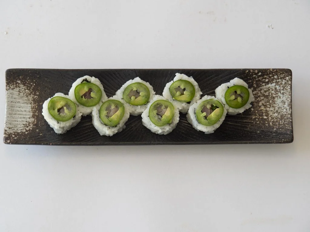 Top view of a black rectangular plate with eight pieces of sushi, each topped with a ring of green jalapeño pepper.