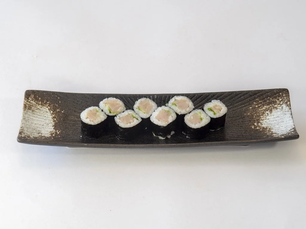 Eight pieces of sushi with raw fish, rice, and avocado on a long, rectangular, dark brown ceramic plate.