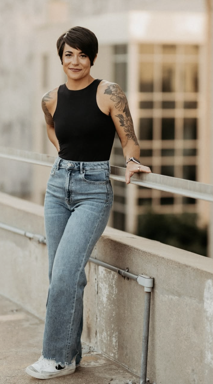 Woman with short dark hair, tattoos on arms, wearing a sleeveless black top, light blue jeans, and white sneakers, standing on a rooftop or balcony with a cityscape in the background.