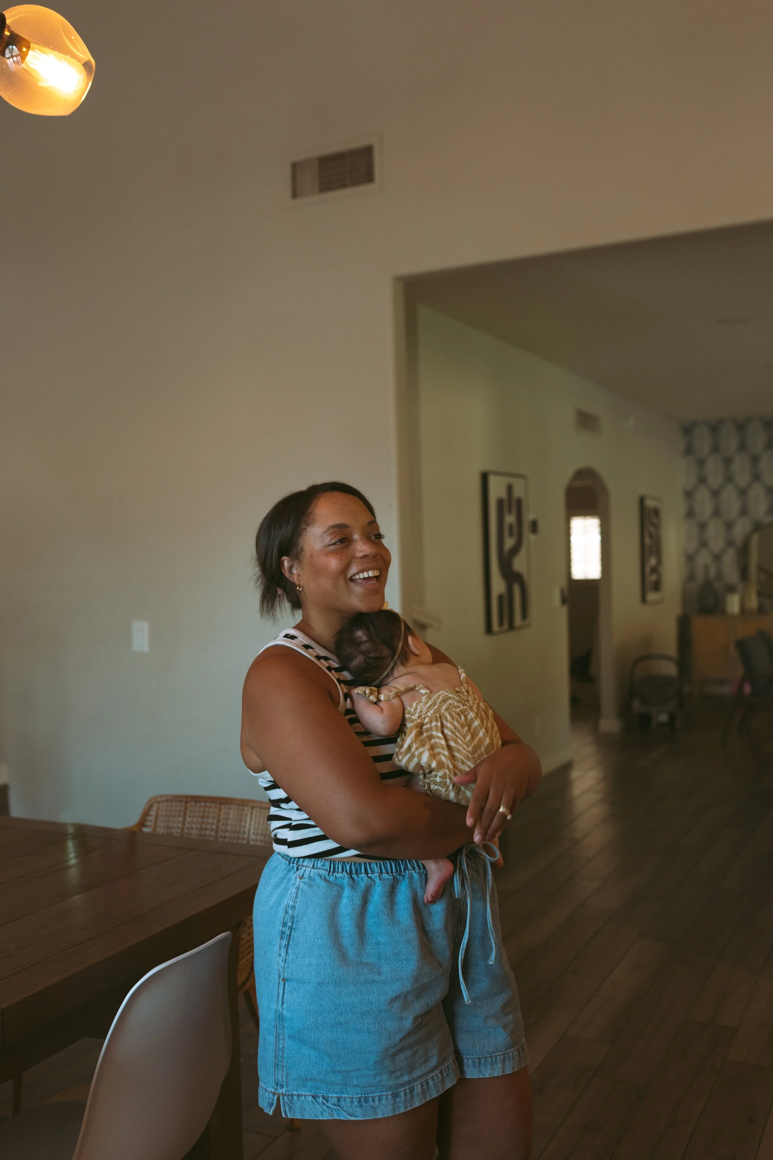 A smiling woman holding a sleeping baby inside a living room with hardwood floors, a dining table, chairs, and framed art on the walls.