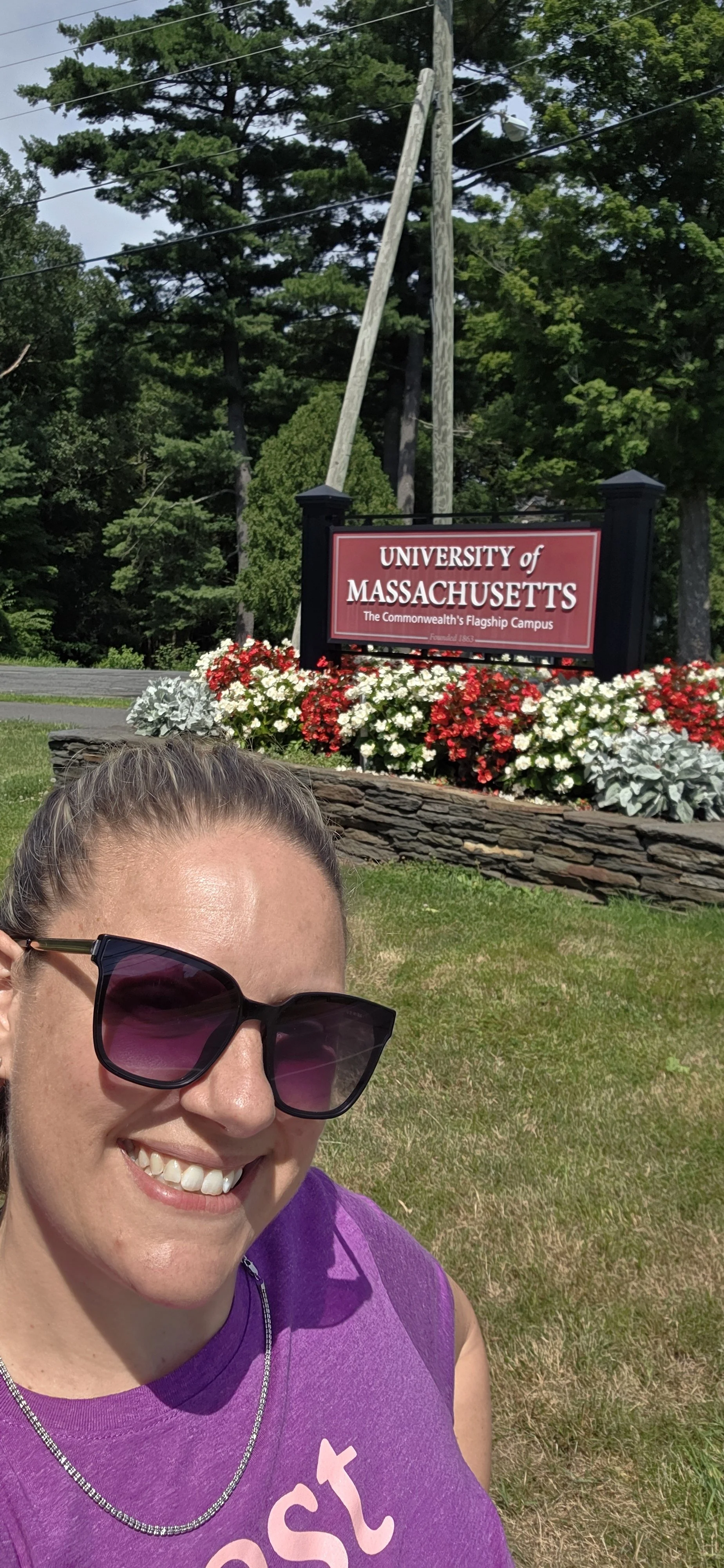 Smiling woman wearing sunglasses and a purple shirt taking a selfie in front of the University of Massachusetts campus sign surrounded by flowers and trees.