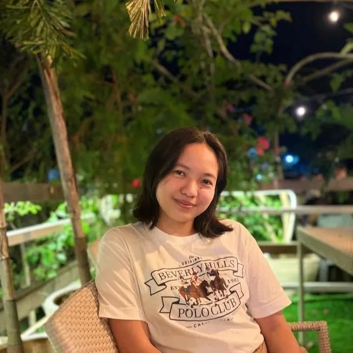 A young woman with black hair sits outdoors at night, smiling softly, surrounded by greenery and string lights, wearing a beige t-shirt with a graphic design.