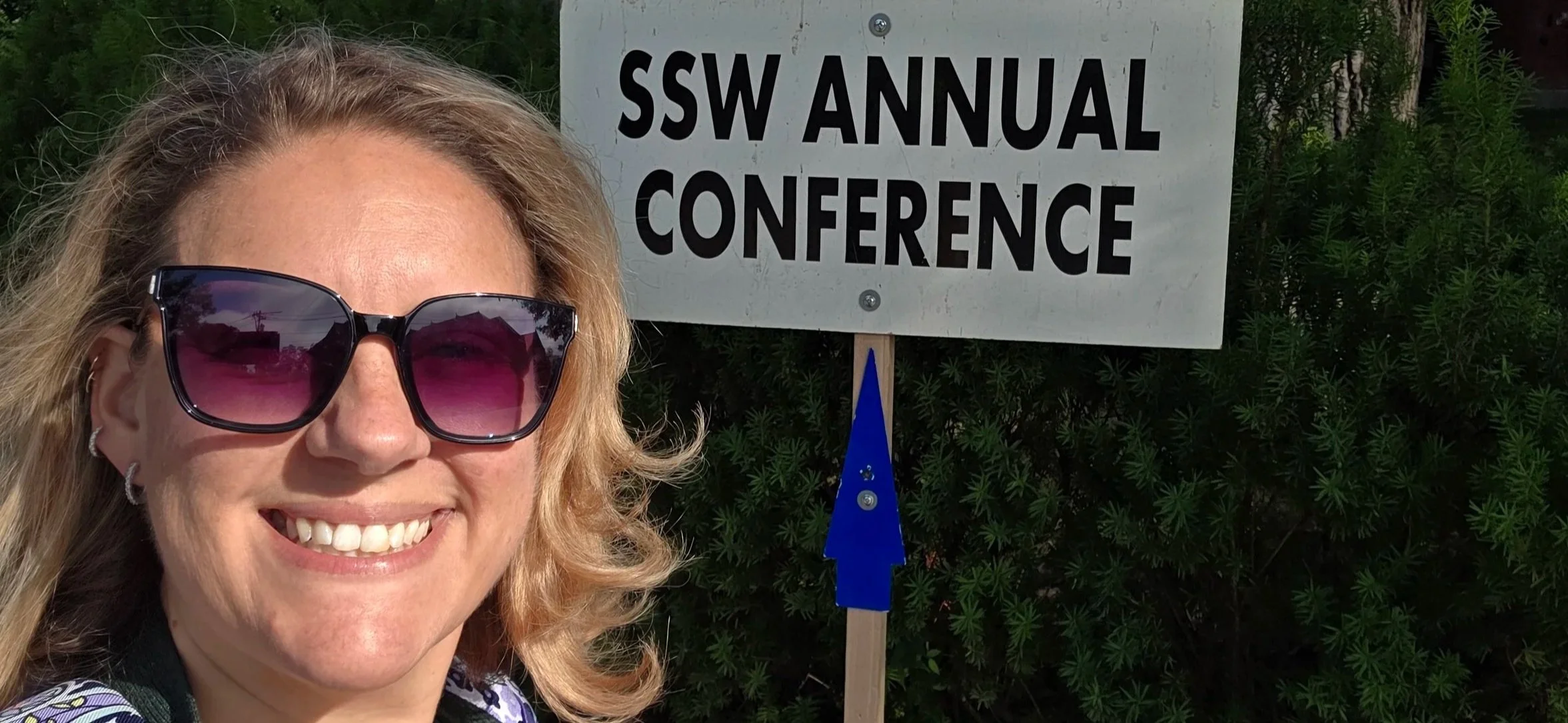 A woman with blonde hair, wearing sunglasses and earrings, smiling, standing outdoors next to a sign that reads 'SSW Annual Conference' with greenery in the background.