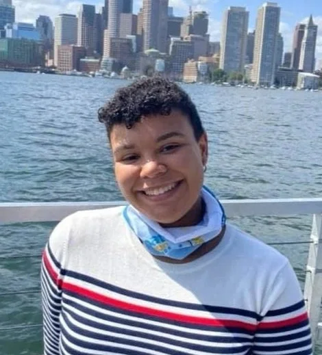 A smiling person standing on a boat with a city skyline in the background.