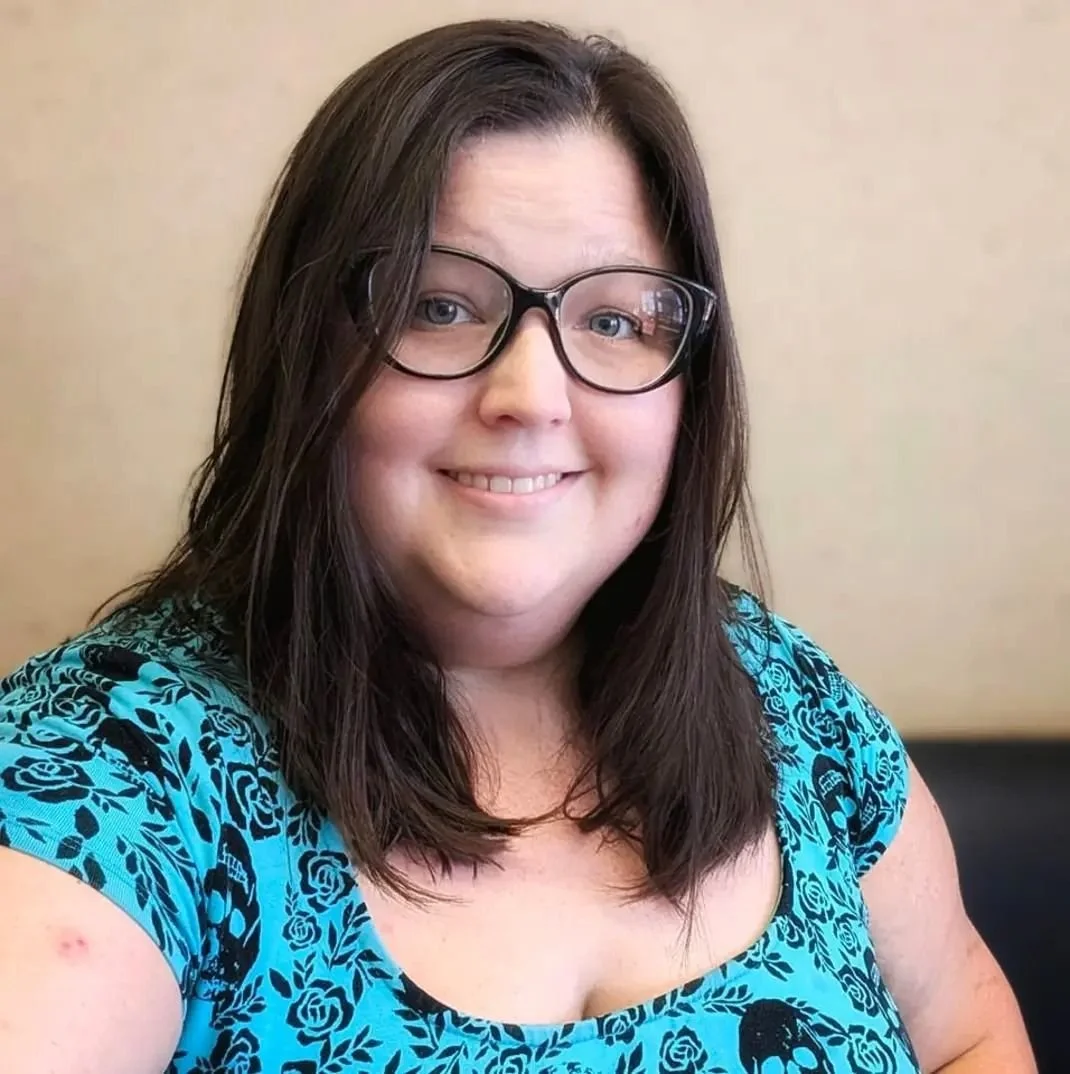 A woman with shoulder-length dark brown hair and glasses, smiling at the camera, wearing a blue patterned top, inside a room with a beige wall background.