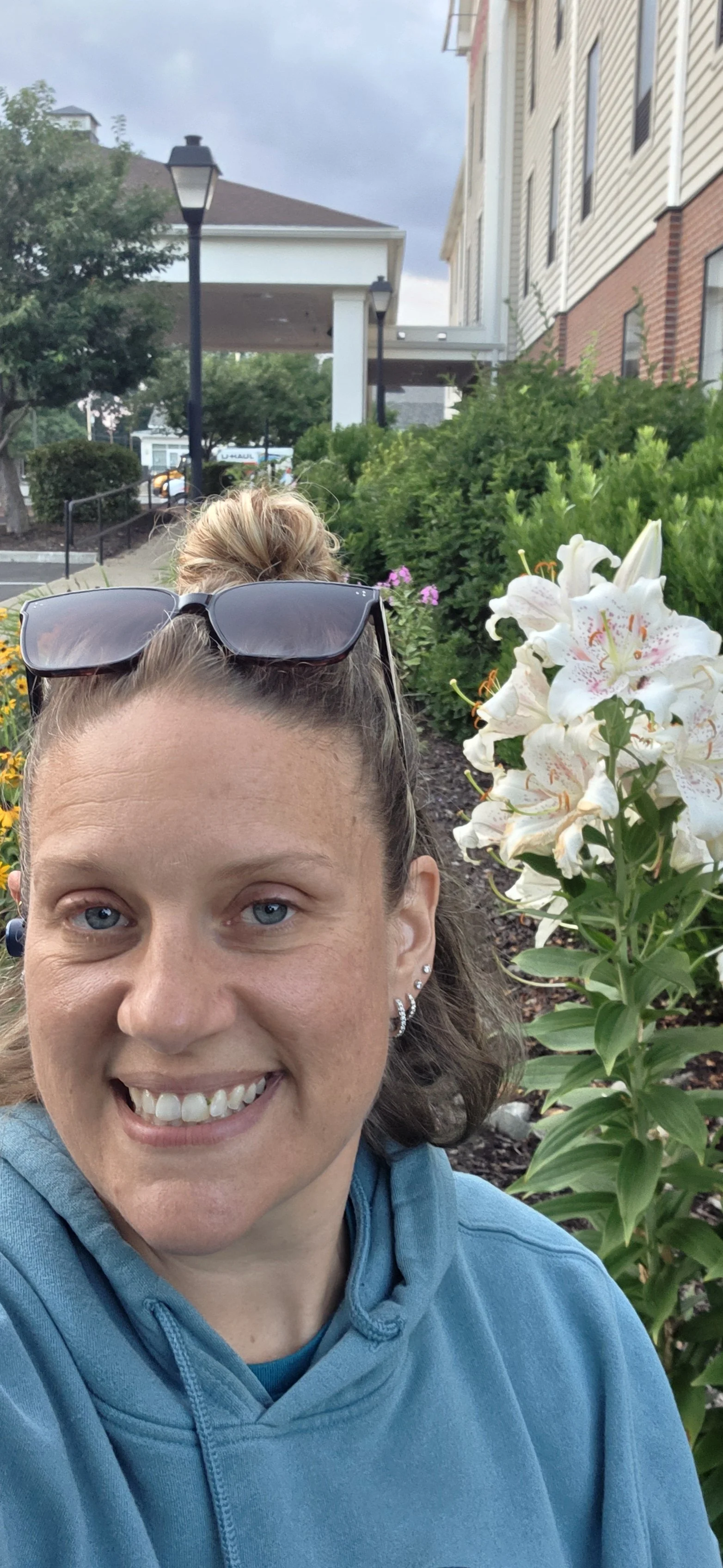 Smiling woman with blue eyes, wearing a blue hoodie and sunglasses on her head, taking a selfie outdoors near white lilies and green shrubs.