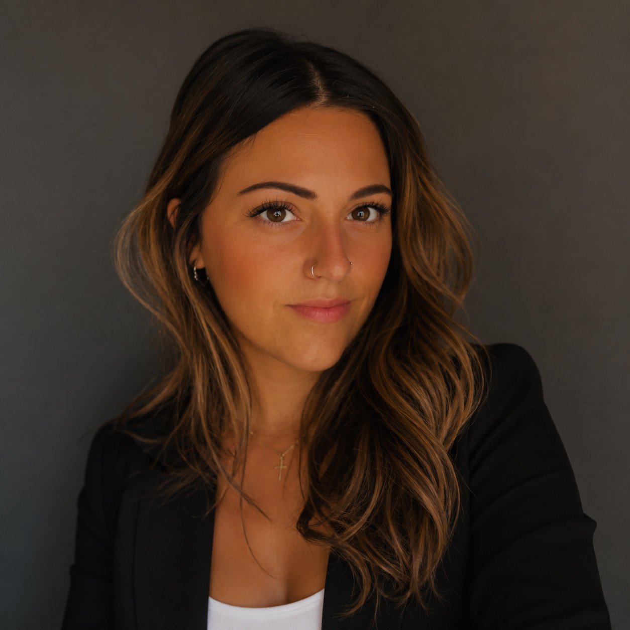 A young woman with brown wavy hair, brown eyes, wearing a black blazer, white top, and jewelry, posing against a dark background.