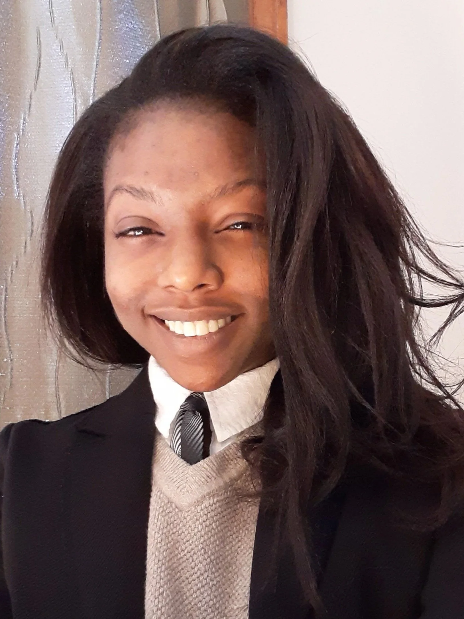 A woman smiling, wearing a black blazer, beige sweater, white shirt, and striped tie, with long dark hair, standing in front of a wooden wall and a white background.