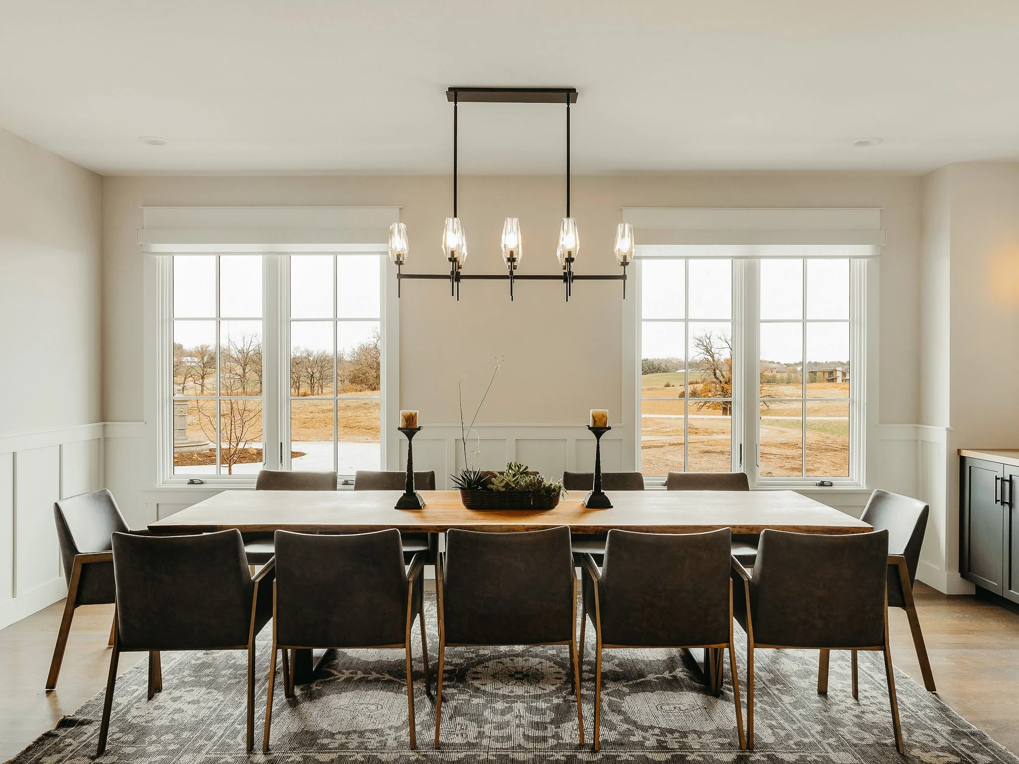 A modern dining room with a large wooden table, eight chairs, and a chandelier hanging above.