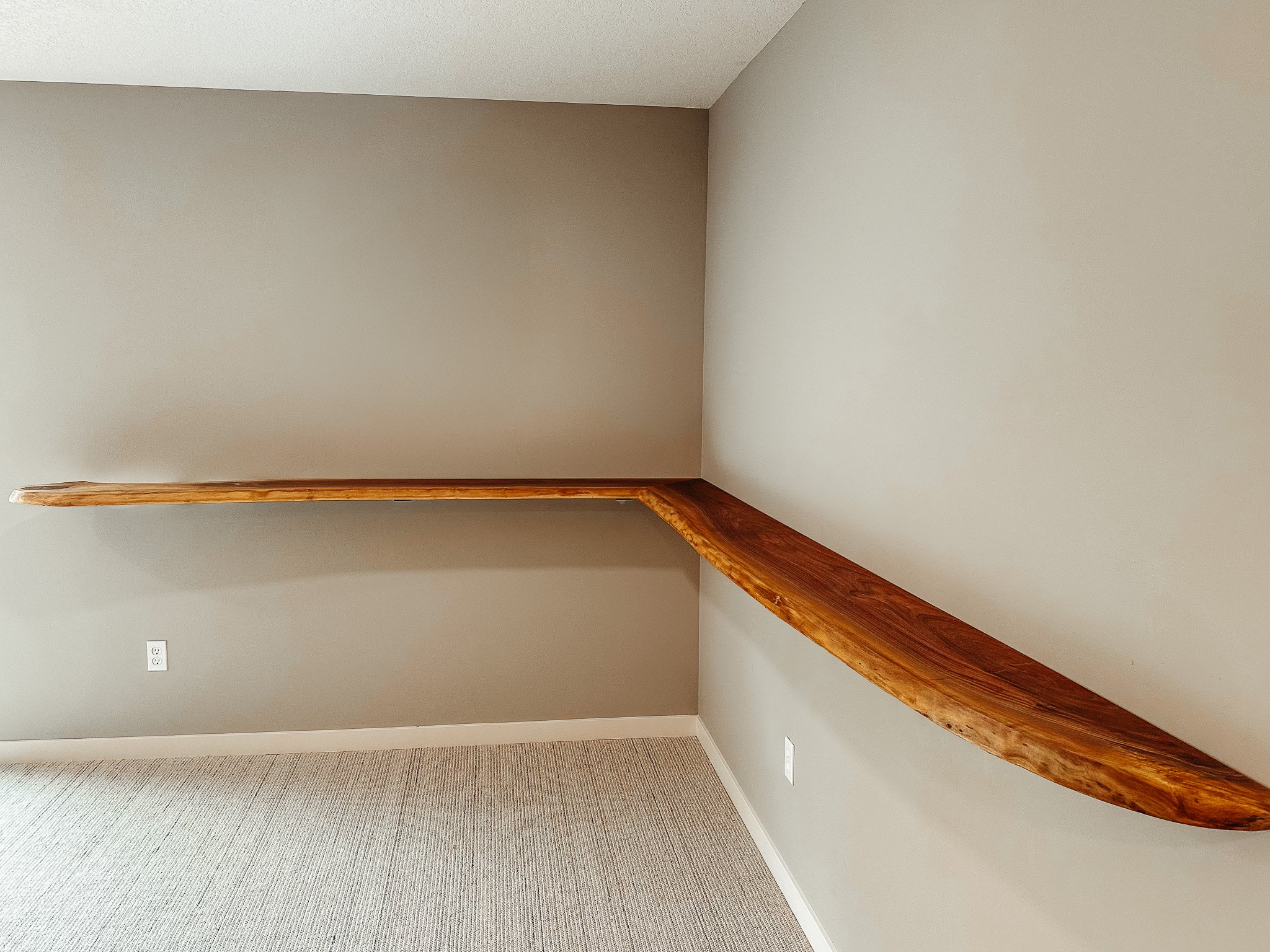 Interior view of a room with a beige wall, a wooden staircase railing, and beige carpeted flooring.