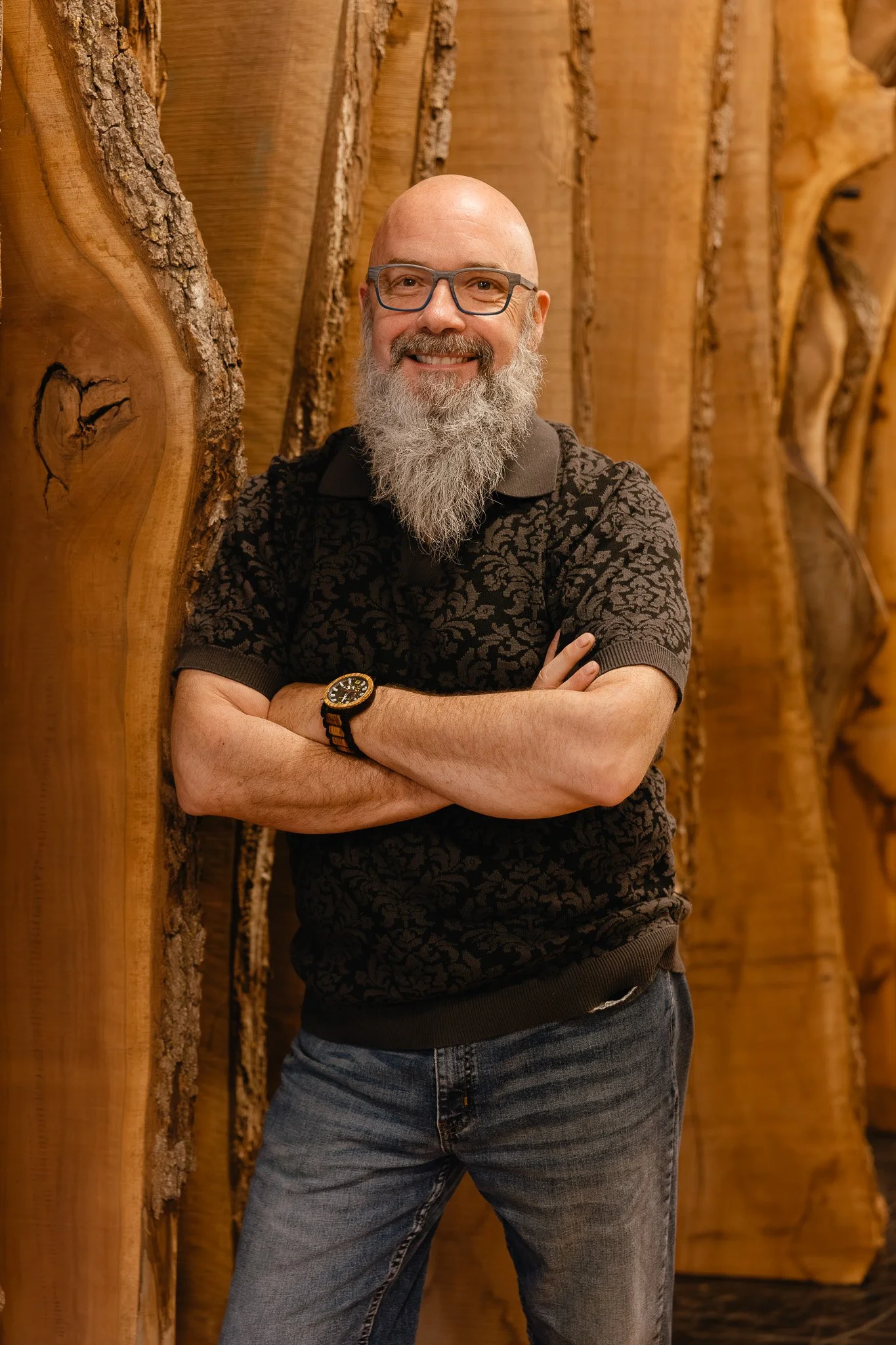 A man with a bald head and a long gray beard is standing with his arms crossed, smiling, in front of a wooden wall.