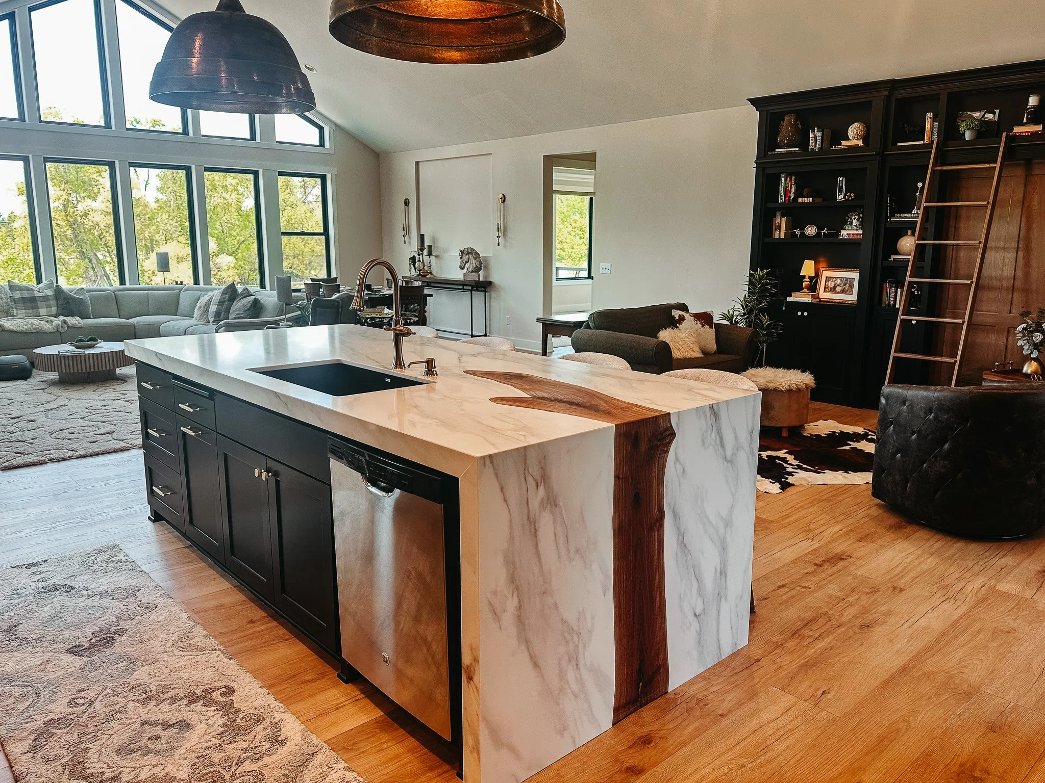 Open-concept living room and kitchen with large windows, a white marble kitchen island, dark cabinetry, a ceiling with two large metallic pendant lights, a gray sectional sofa with pillows, a black bookshelf with decorations and ladder, and hardwood 