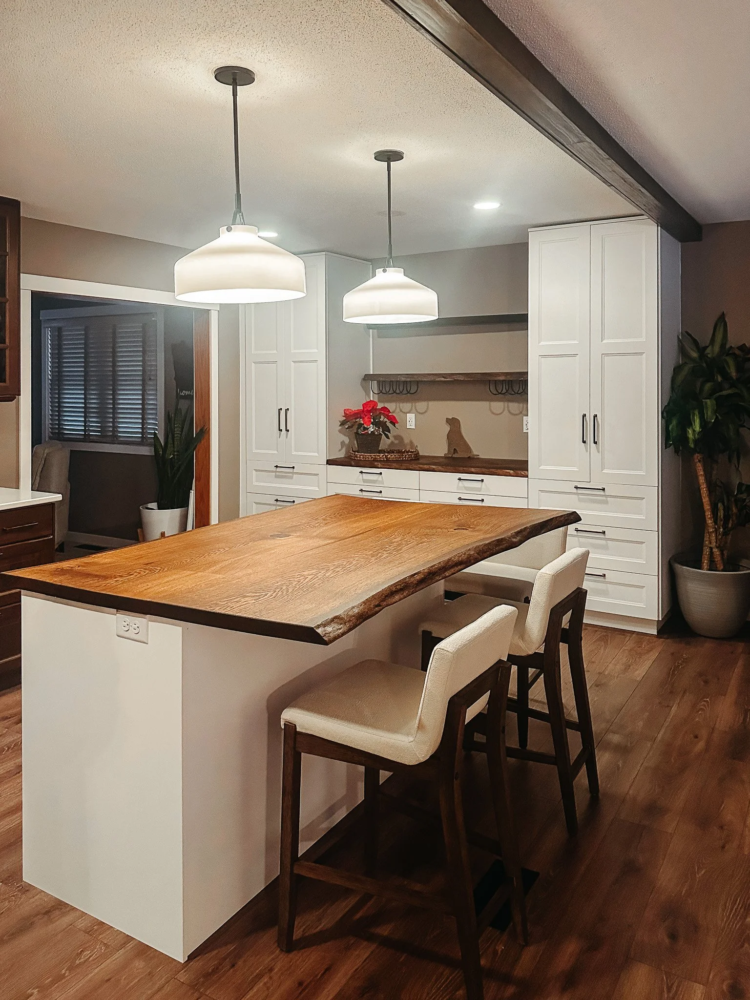 A modern kitchen with white cabinets, a wooden island, and pendant lights hanging from the ceiling. There is a plant in a white pot and holiday decorations on the countertop.