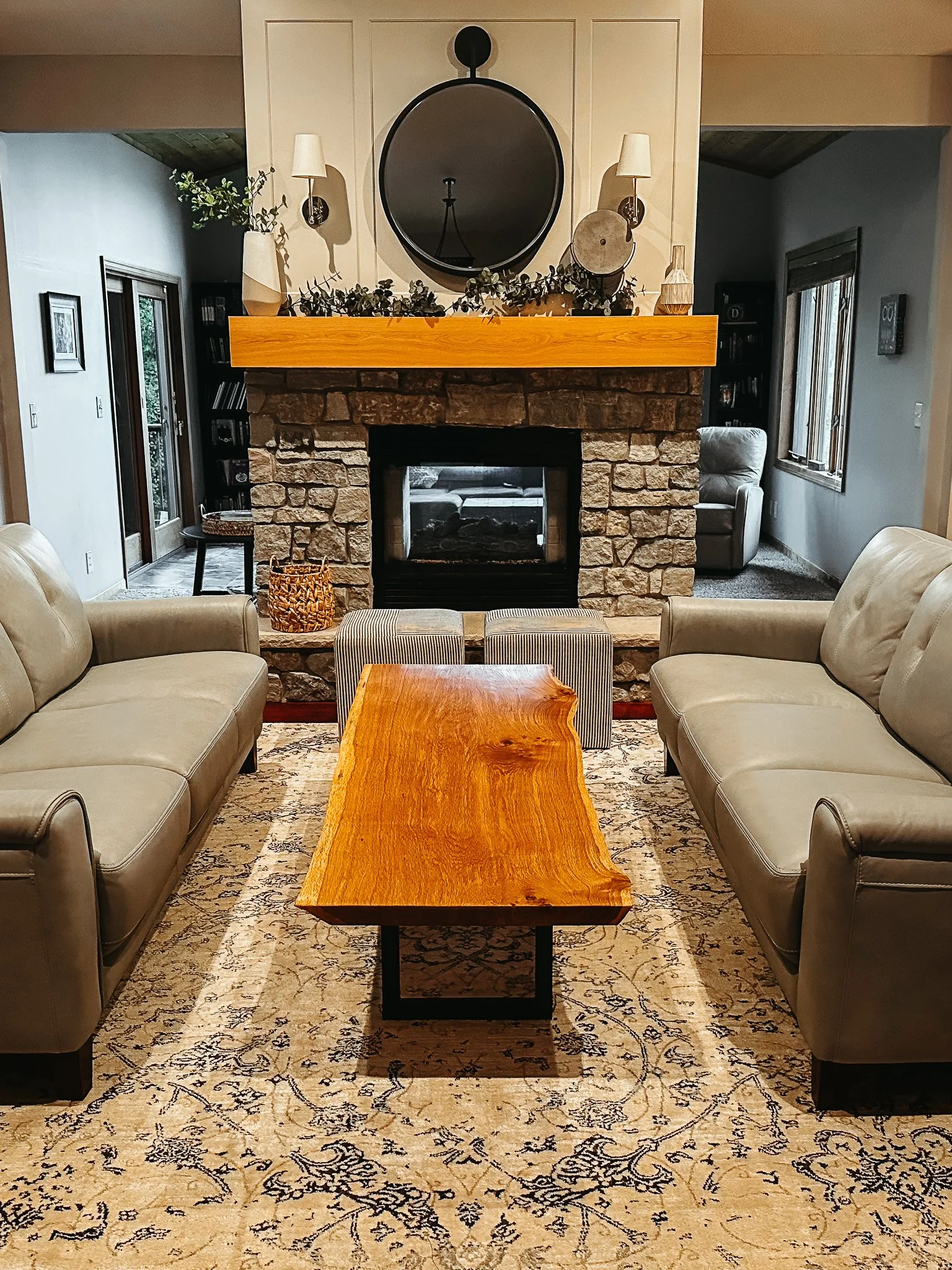 A living room featuring a stone fireplace with a wooden mantel, surrounded by beige leather sofas, a patterned rug, a wooden coffee table, and decorative items including a mirror, vases, and wall-mounted lamps.