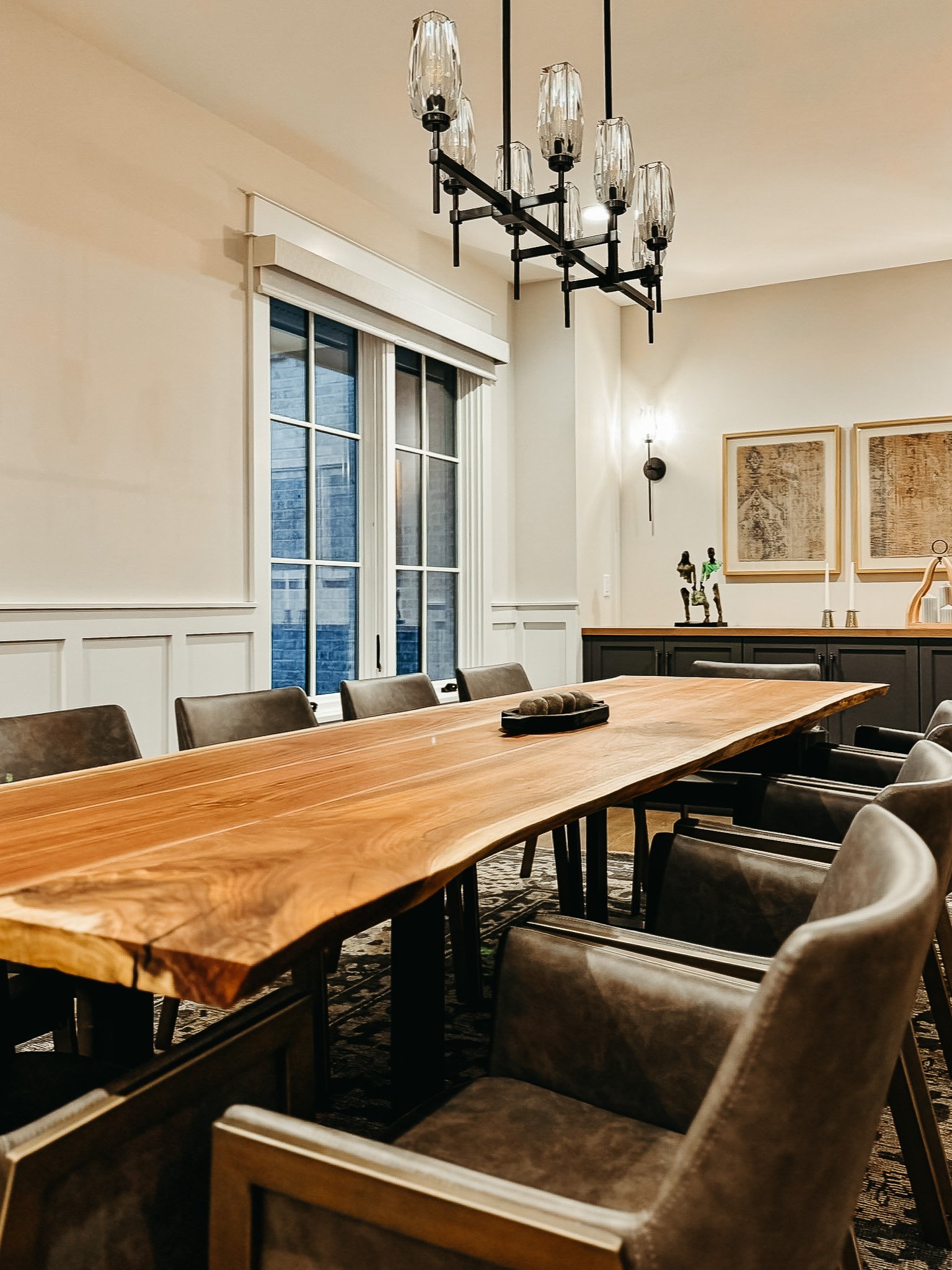 A dining room with a large wooden table, surrounded by cushioned chairs, a chandelier hanging from the ceiling, and artwork on the wall.